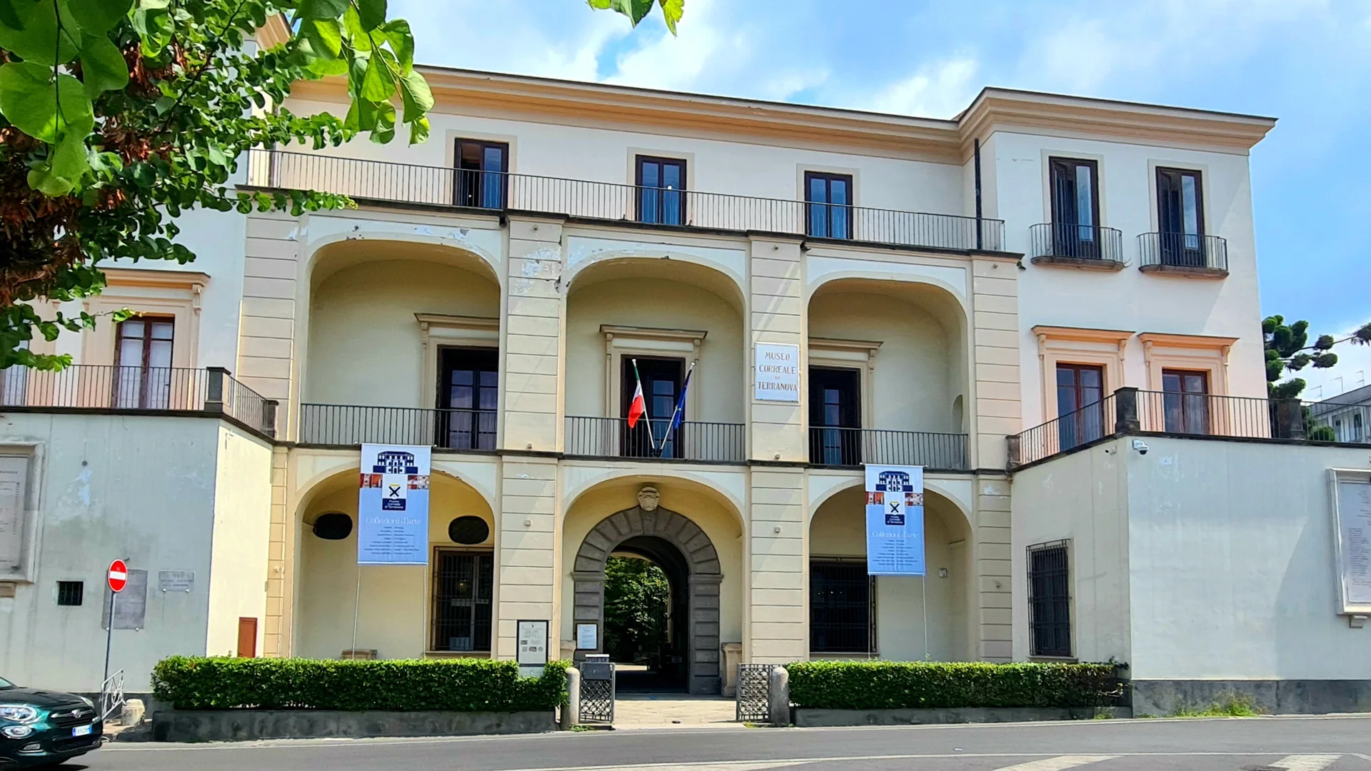 Die cremefarbene Fassade des Museo Correale di Terranova in Sorrent, mit drei bogenförmigen Loggien über einem steinernen Torbogen, Balkonen mit Eisengeländern, einer Trikolore am Eingang und einem belaubten Zweig in der Ecke.