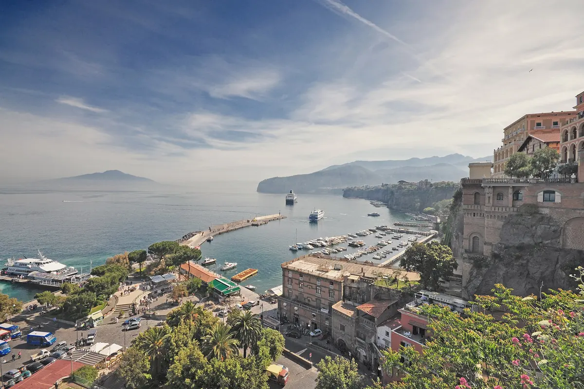 Ein Blick über den Hafen von Sorrento mit einem langen Pier, kleinen Booten und Klippen auf der rechten Seite sowie der schemenhaften Silhouette des Vesuvs jenseits des Wassers unter einem wolkenverhangenen Himmel.