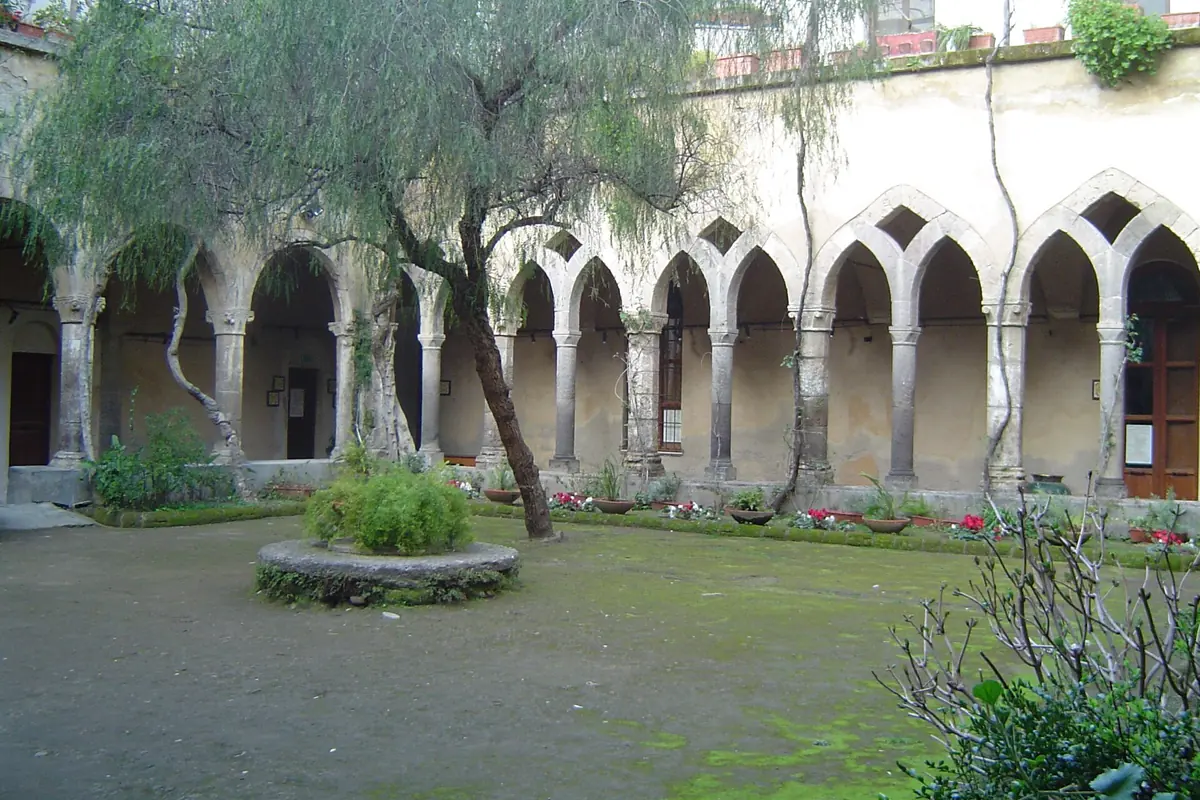 Ein Kreuzganggarten in Sorrento mit einer Reihe spitzbogiger Arkaden auf schlanken Säulen und einem runden Steinbrunnen im Gras.