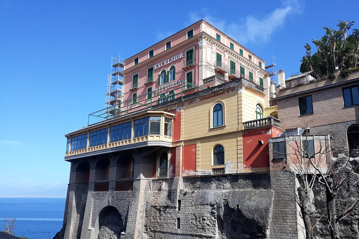 Ein rosa- und ockerfarbenes Hotel mit grünen Fensterläden und einem verglasten Balkon auf einer hohen Steinklippe über dem Meer in Sorrento.