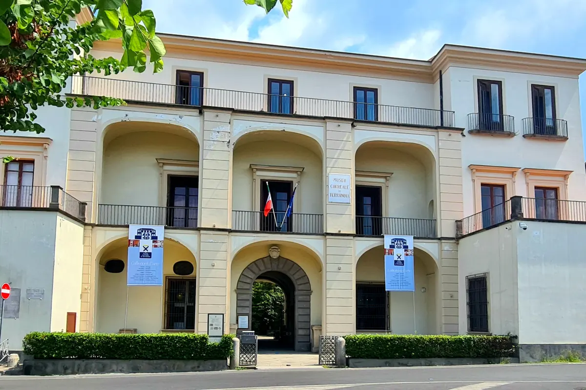 Krémově zbarvené průčelí muzea Museo Correale di Terranova v Sorrentu se třemi obloukovými lodžiemi nad kamennou branou, balkony se železným zábradlím a zelenou větví v rohu.