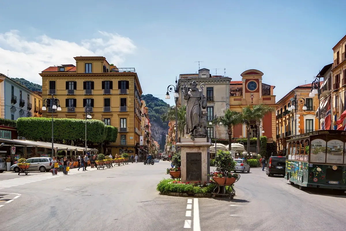 Ein weiter Platz in Sorrento mit ocker- und terrakottafarbenen Gebäuden, einer Statue auf einem Sockel in der Mitte und Palmen.