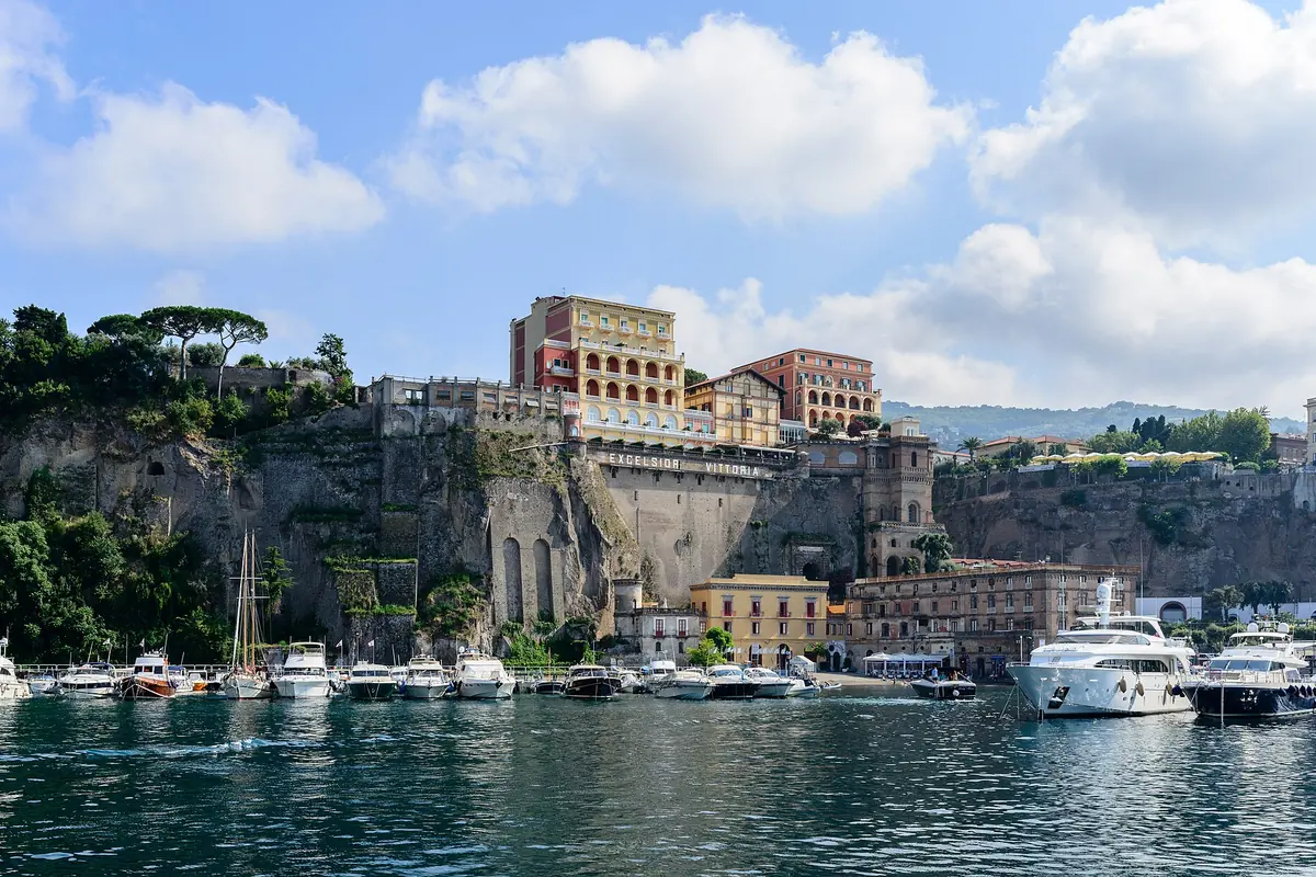 Vertäute Boote und eine weiße Yacht im Hafen von Sorrento unter einer hohen Klippe, gekrönt von rosa- und ockerfarbenen Gebäuden.