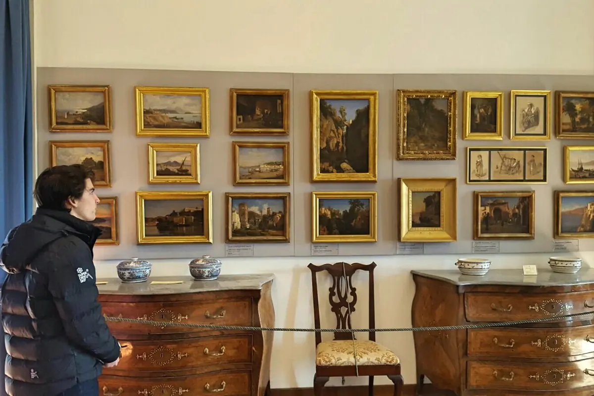 Ein Museumsraum mit einer Wand voller kleiner, goldgerahmter Landschaftsgemälde über zwei dunklen Holzkommoden und einem Stuhl.